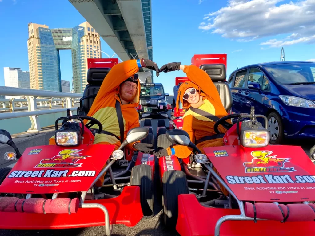 Two people in orange costumes form a heart with their arms while sitting in red Street Kart go-karts on a sunny city street under a bridge.