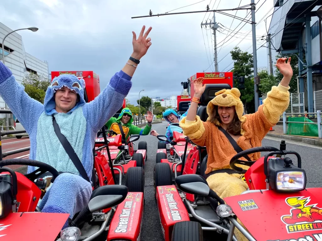 Group of friends in blue and brown bear onesies ride red Street Kart go-karts, waving at the camera on a city street.